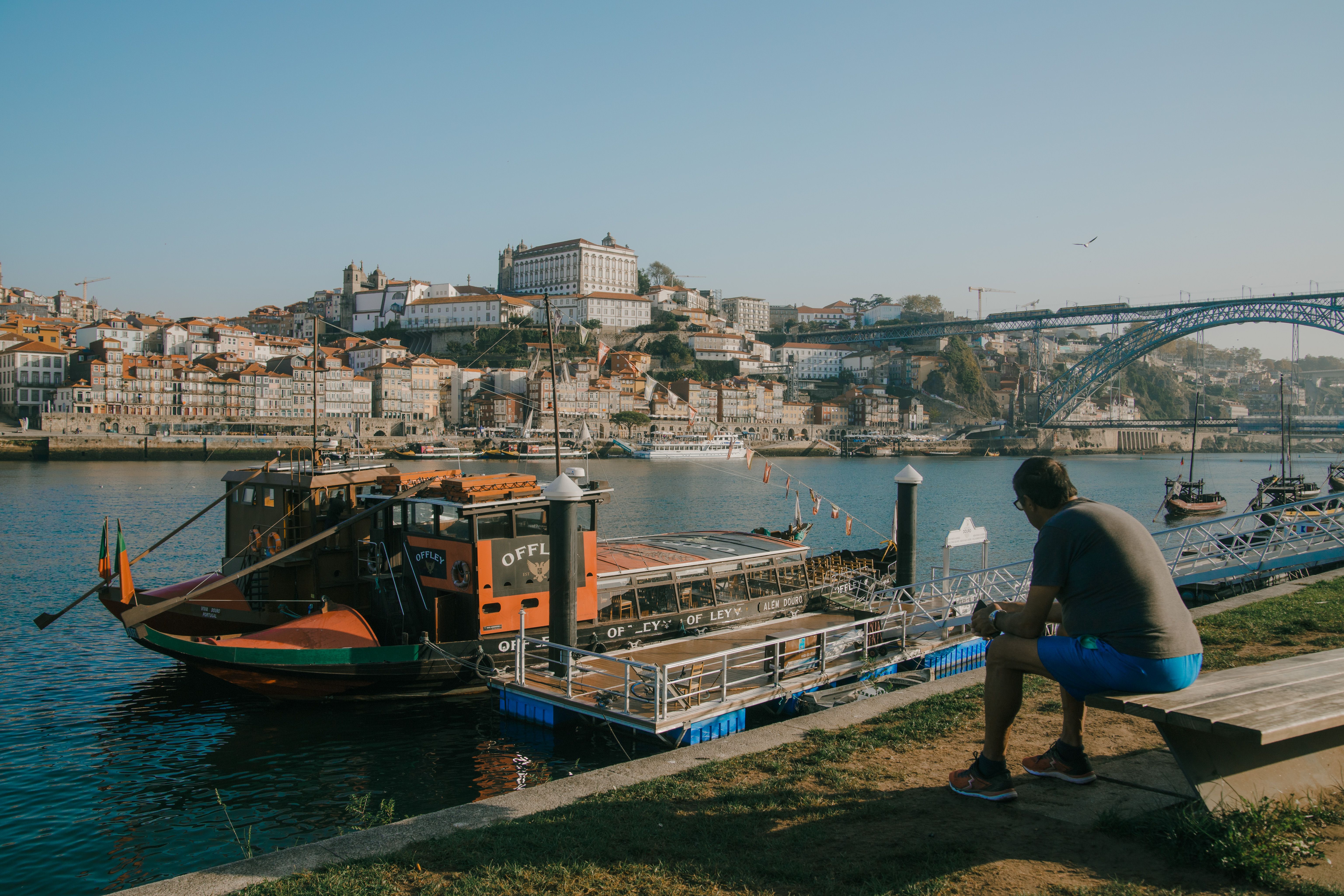 A man sits by the river in Porto, overlooking boats and cityscape, with an iconic bridge in view.