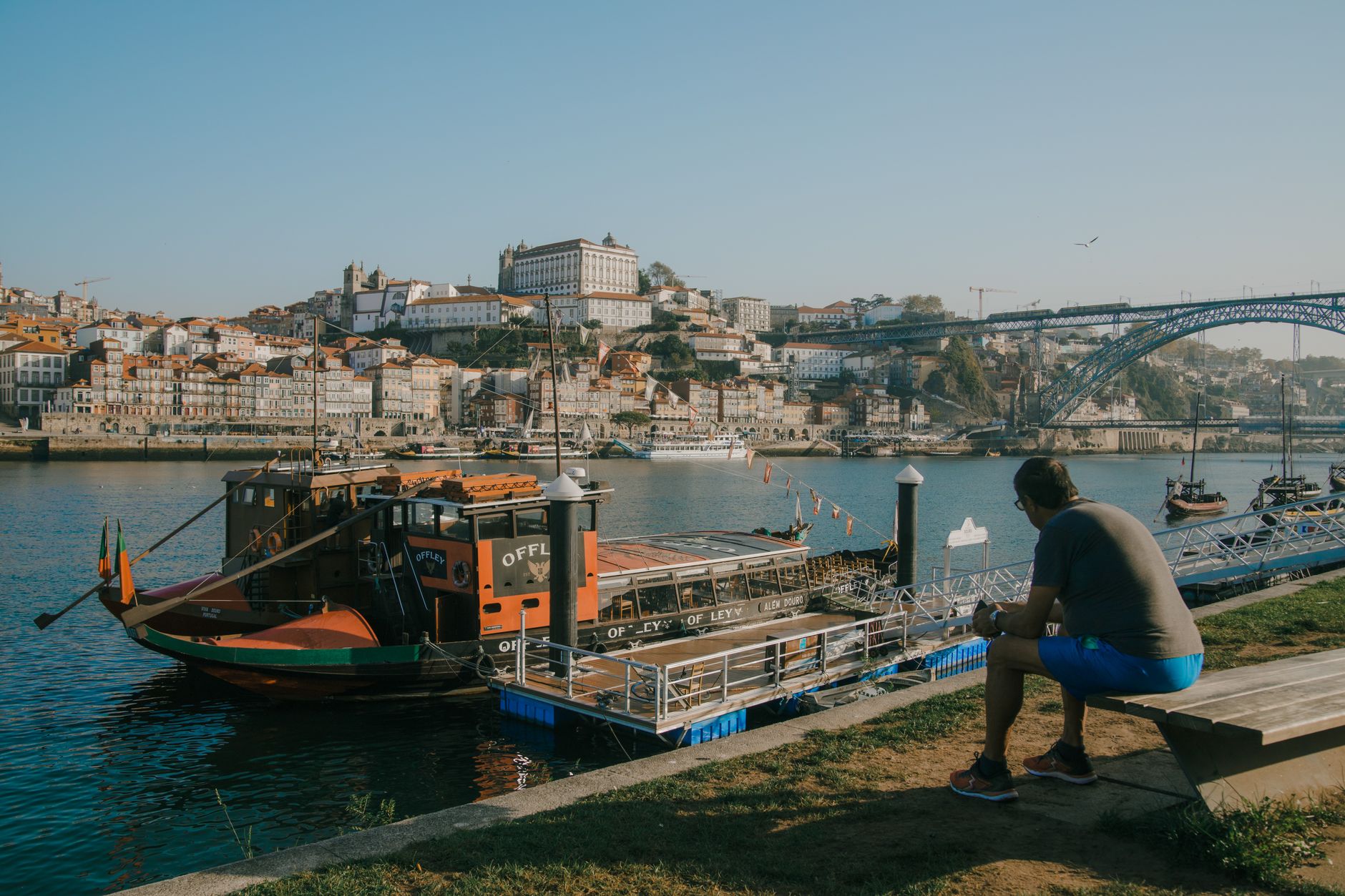 A man sits by the river in Porto, overlooking boats and cityscape, with an iconic bridge in view.
