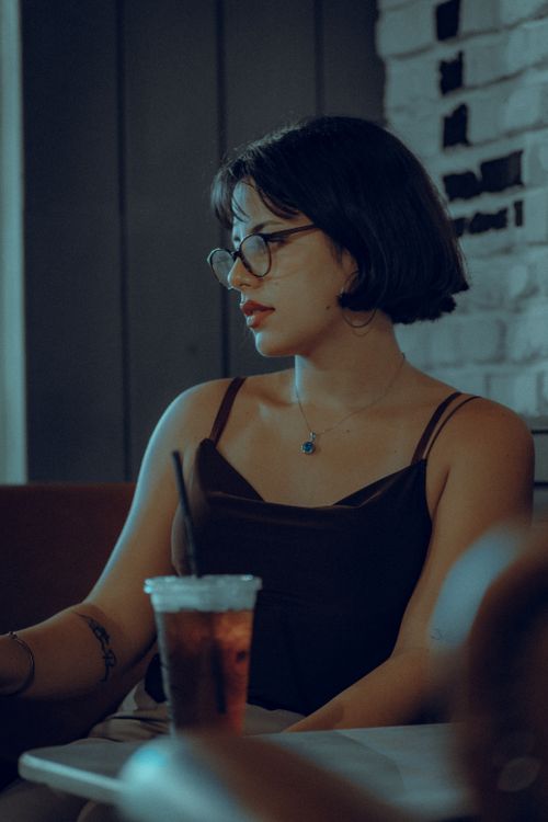 Free Young woman wearing glasses, sitting in a cafe with a drink, looking calm and relaxed. Stock Photo