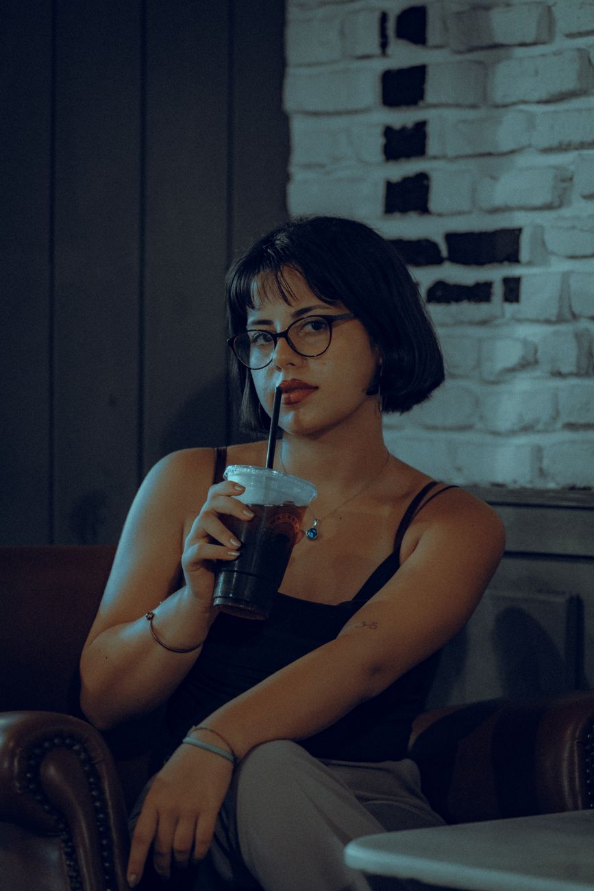 A stylish woman with eyeglasses sipping a drink in a cozy café setting.