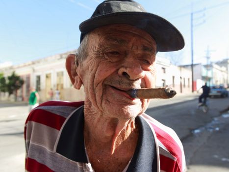 A senior man in a cap smokes a cigar, enjoying a sunny day on a vibrant street.
