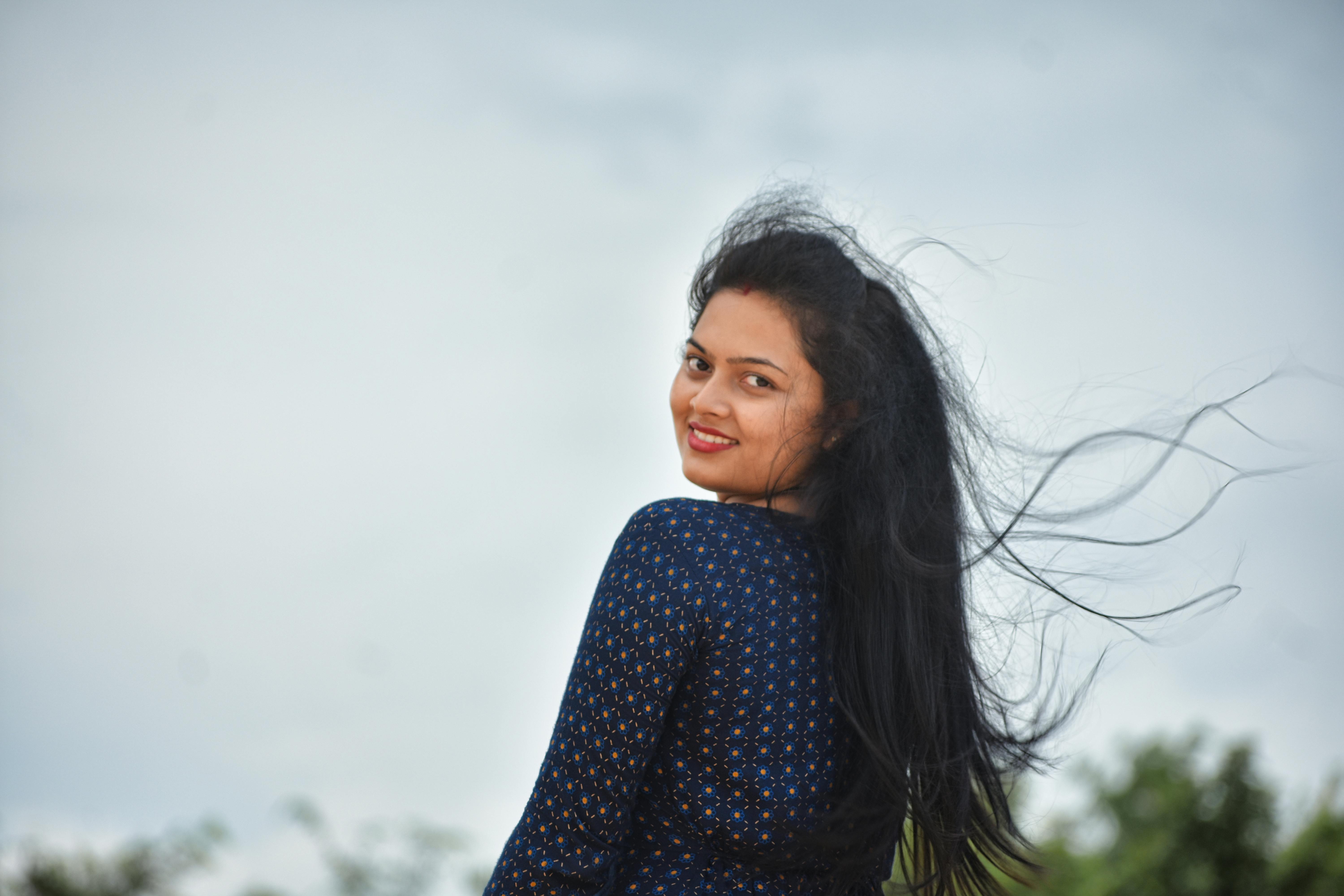 A woman in a blue top with long hair smiles at the camera, set against a clear sky outdoors.