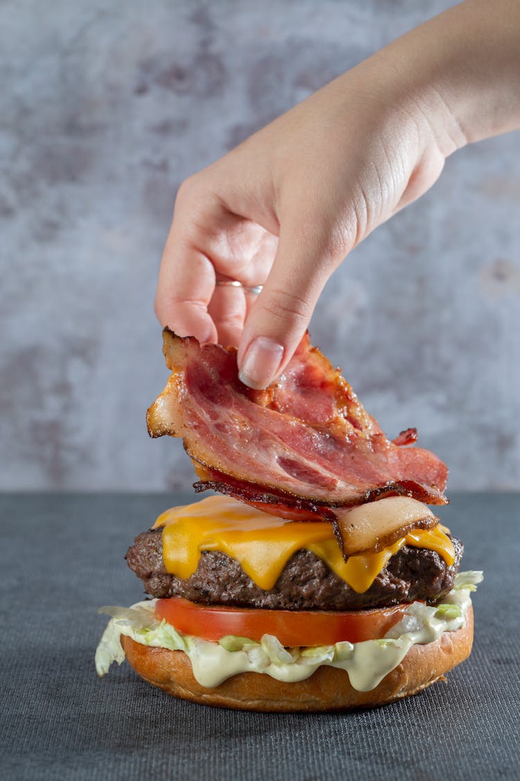 Person Putting Bacon On A Burger 