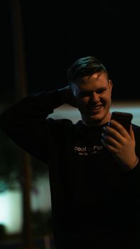 A young caucasian man smiling while using his smartphone outdoors at night.