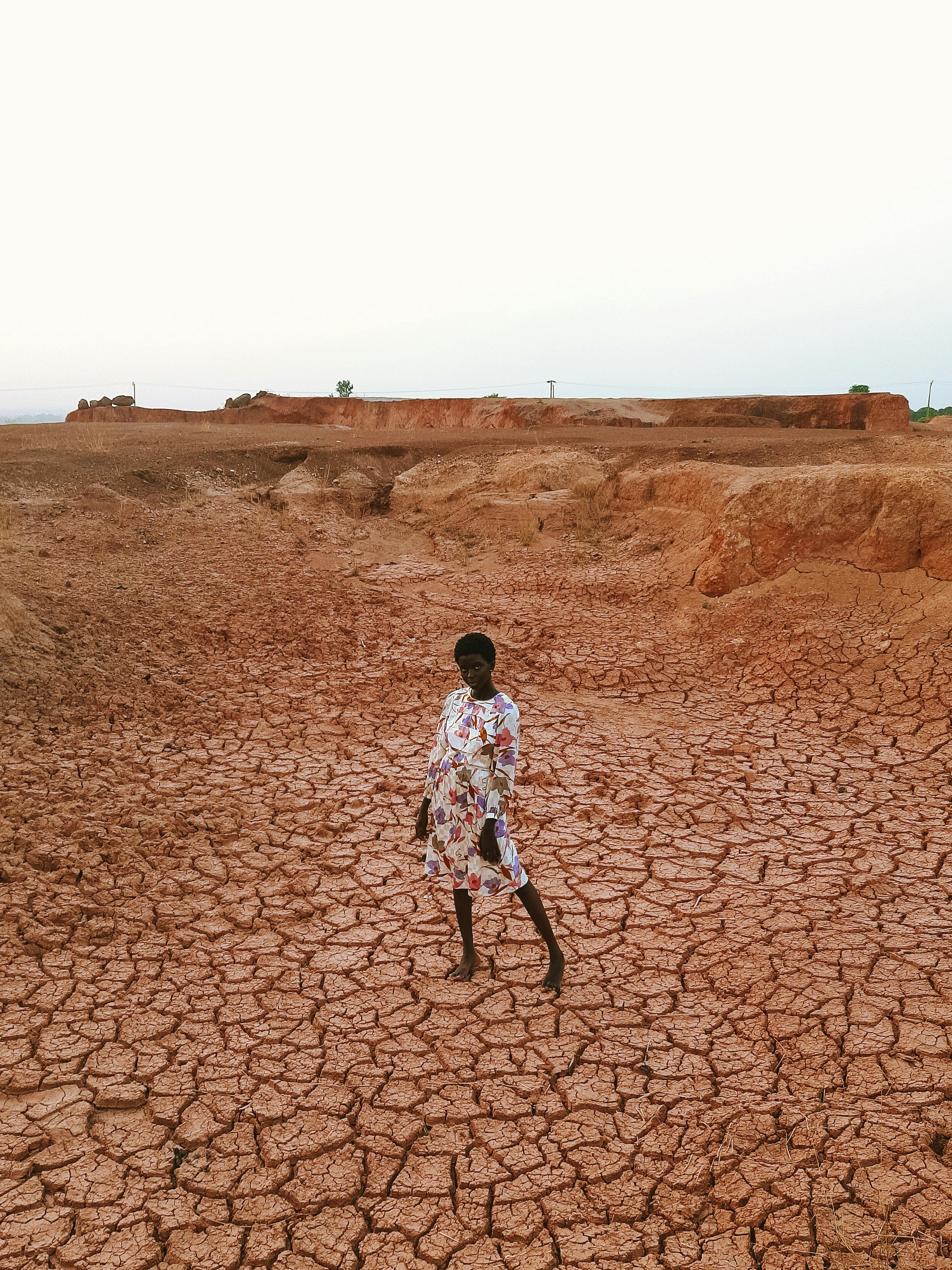 A woman in a floral dress stands on cracked, arid soil, symbolizing resilience in a barren landscape.