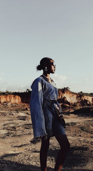 A fashionable woman in a denim dress stands confidently in a barren wasteland on a clear day.