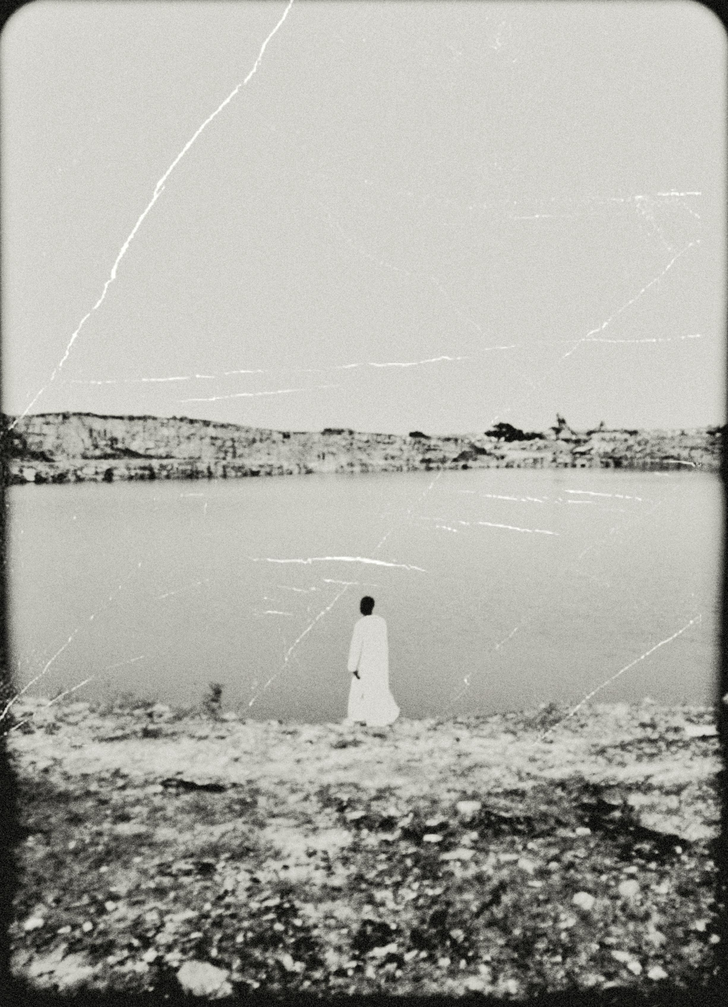 A solitary figure stands by a serene lake in a vintage, monochrome style.