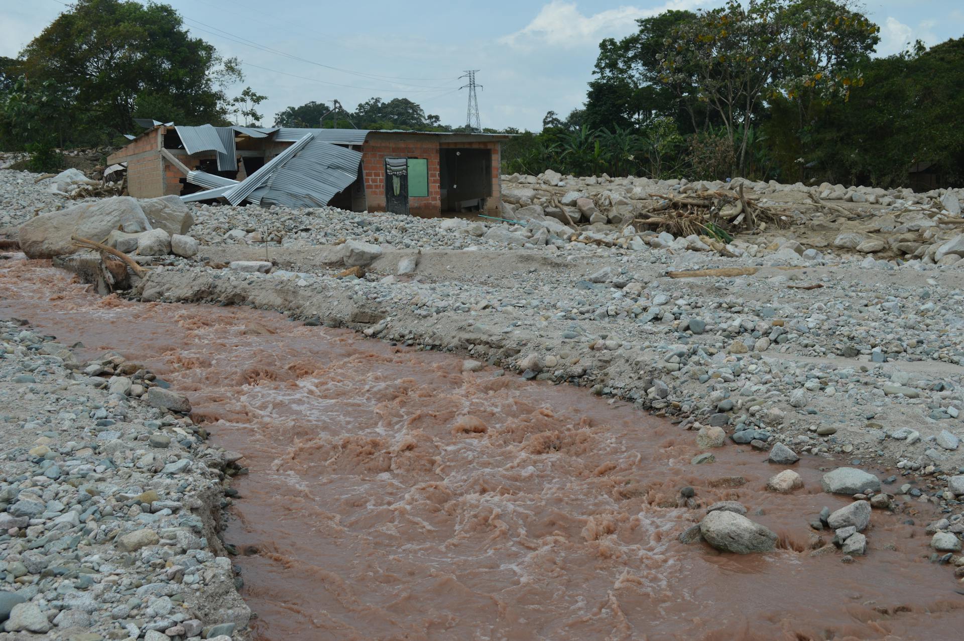 A damaged house stands near a muddy river after a flood, illustrating the impact of natural disasters.