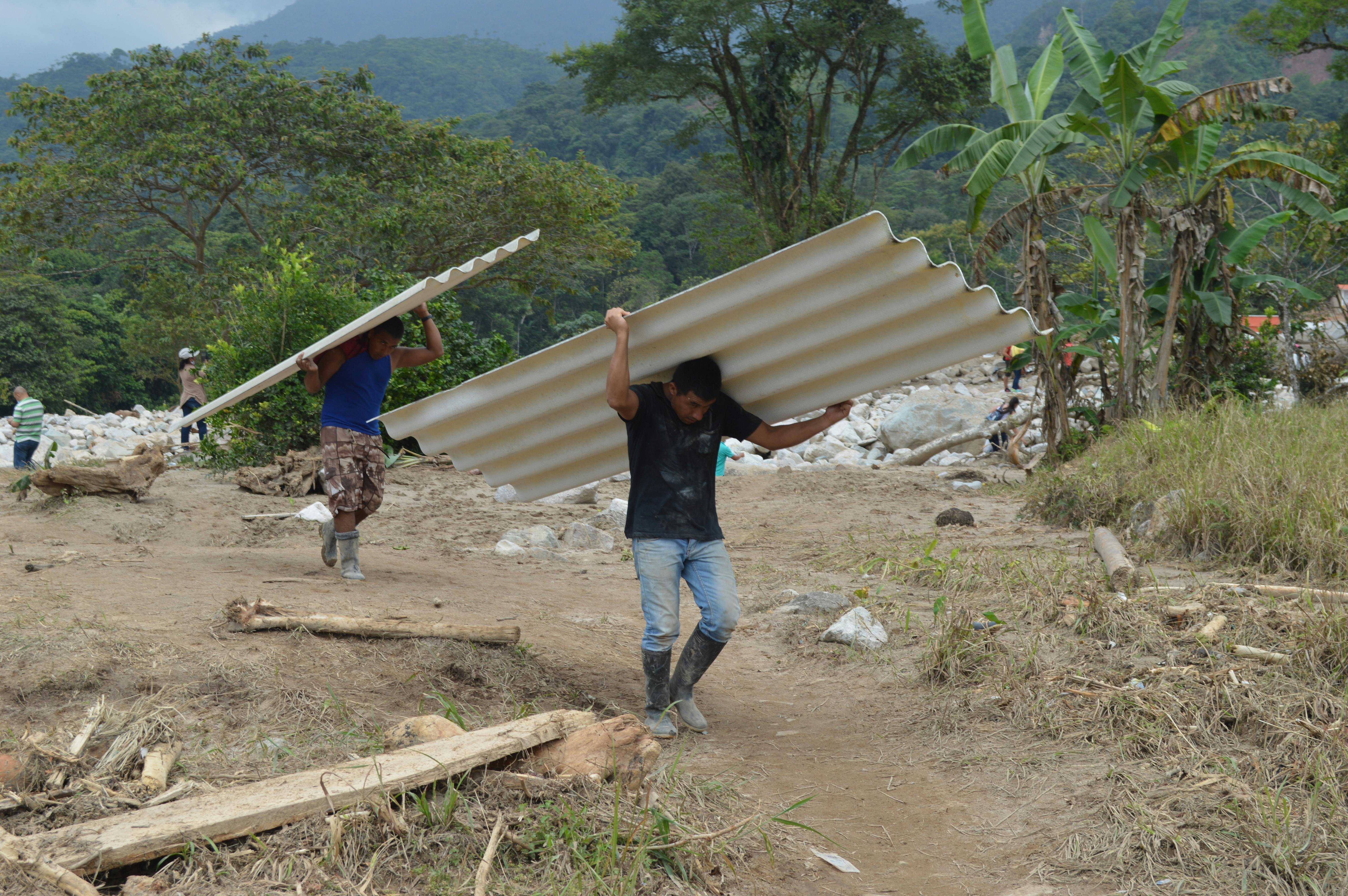 Men carrying roofing sheets at a construction site surrounded by lush forest in Mocoa, Colombia.