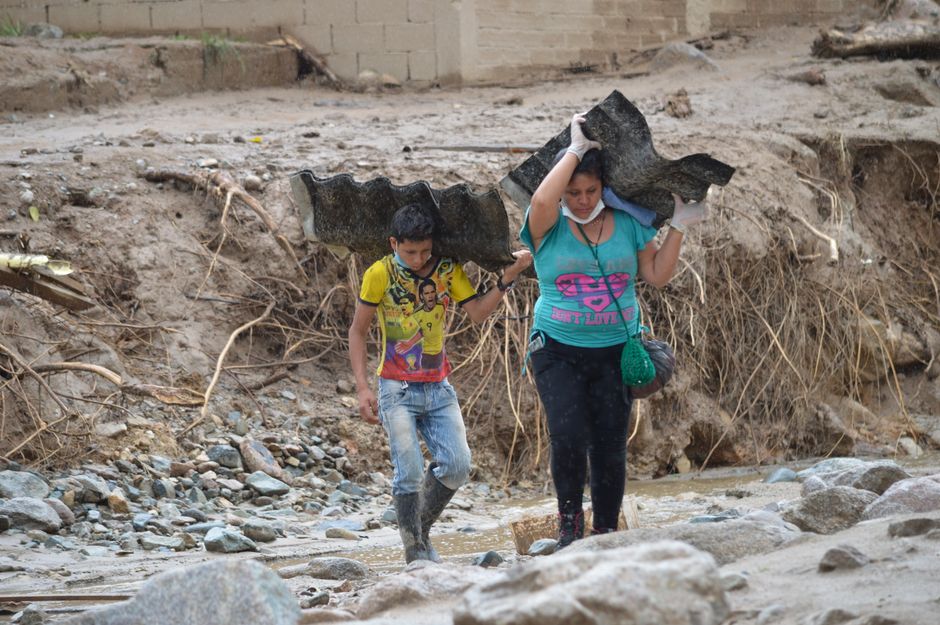 Two individuals carry sheet metal through a rugged terrain in Mocoa, showcasing resilience.
