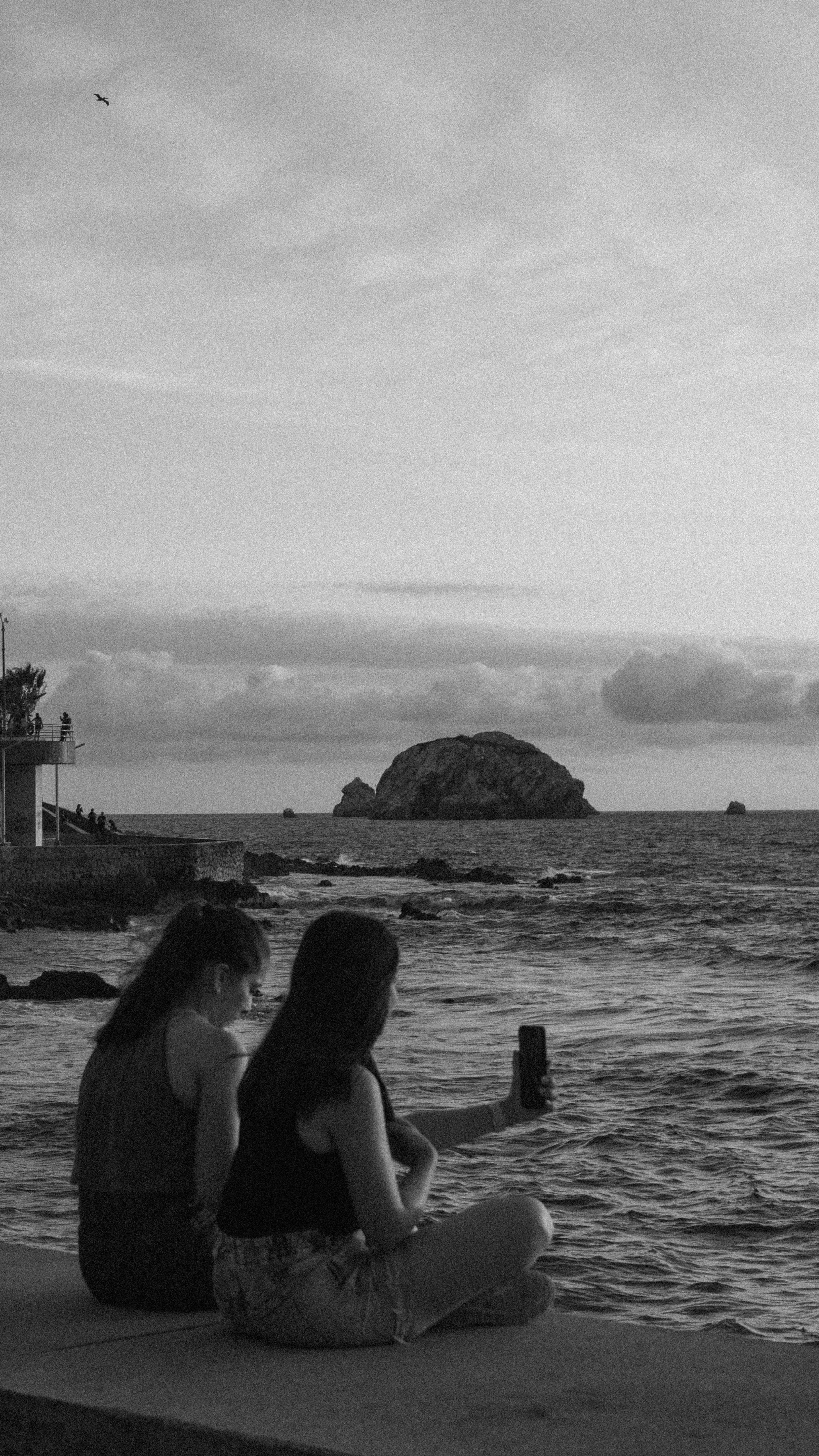 Two women sitting by Mazatlán's sea, capturing the serene sunset with a mobile.