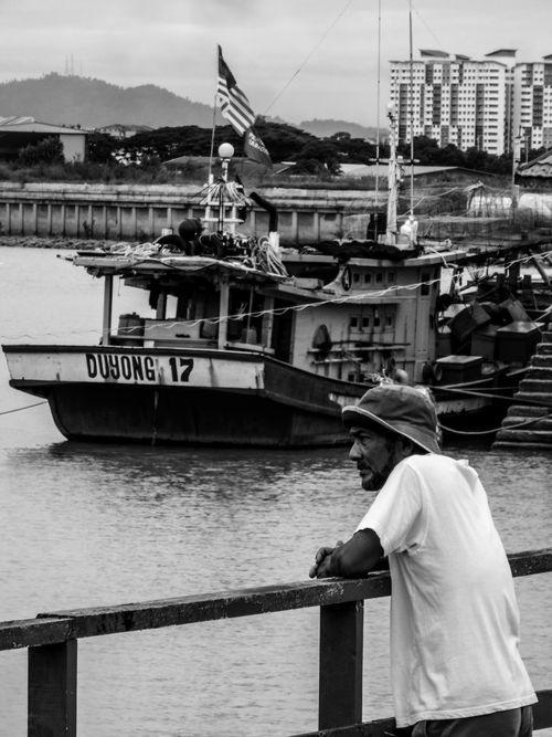 Free An East Asian man leans on a railing by a pier in Malaysia, with a boat in the background. Stock Photo