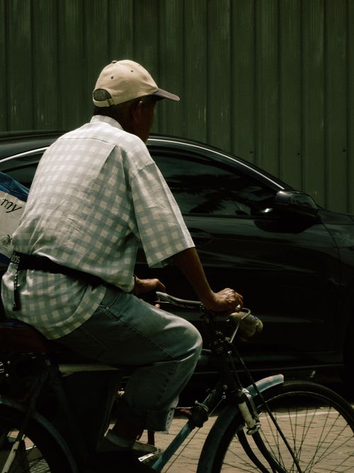 Free A man rides a bicycle past a black car on a sunny day in Malaysia. Stock Photo