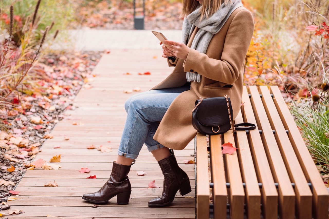 Woman in a camel coat, jeans and brown heeled ankle boots sitting on a park bench