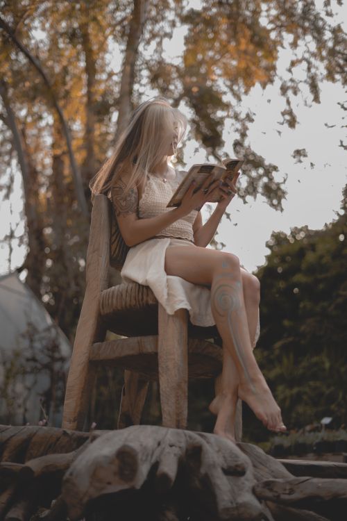 Free A woman sits barefoot on a wooden chair outdoors, reading a book in a peaceful natural environment. Stock Photo