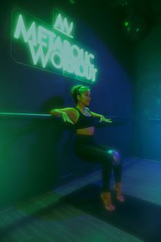 Fit woman in neon gym environment performing a wall sit, highlighting modern fitness culture.