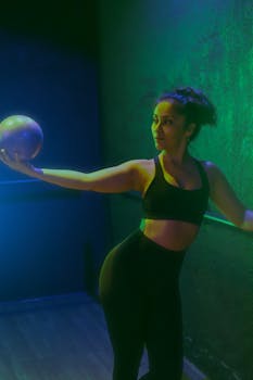 A woman elegantly poses with a gym ball in a dimly lit studio, showcasing strength and balance.
