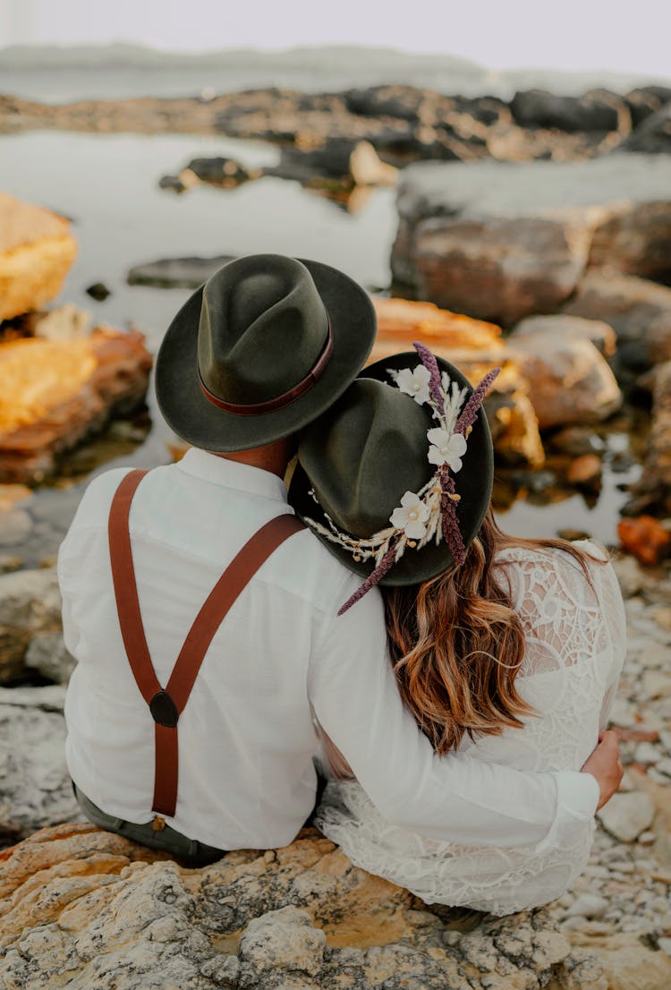 Couple In Hats Sitting On Stones On Beach