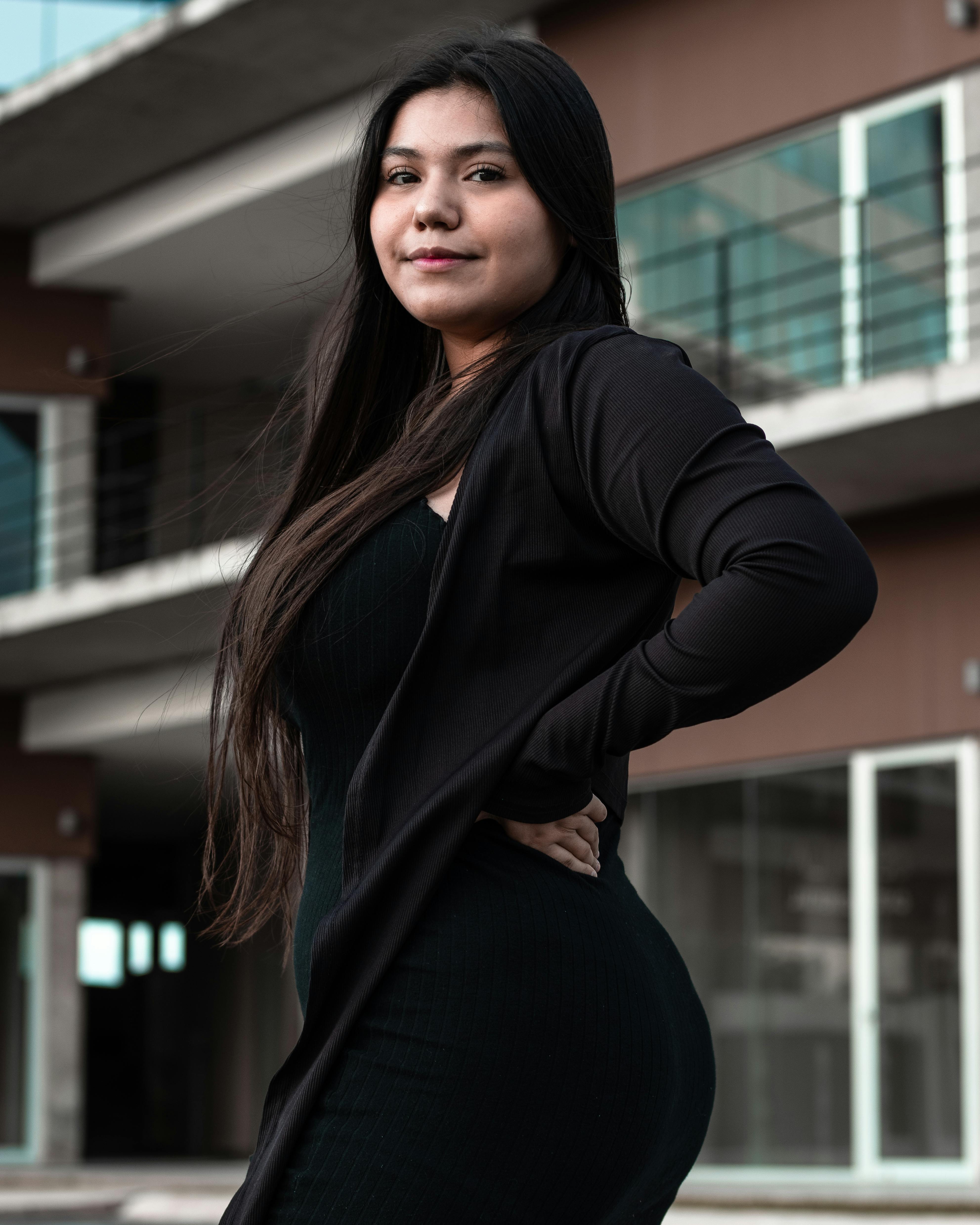 Stylish woman in black posing confidently outdoors against a modern building backdrop.