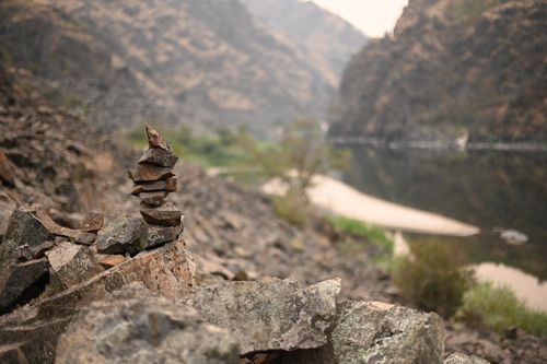 Free A serene rock cairn balanced near a calm river amidst rugged cliffs. Stock Photo