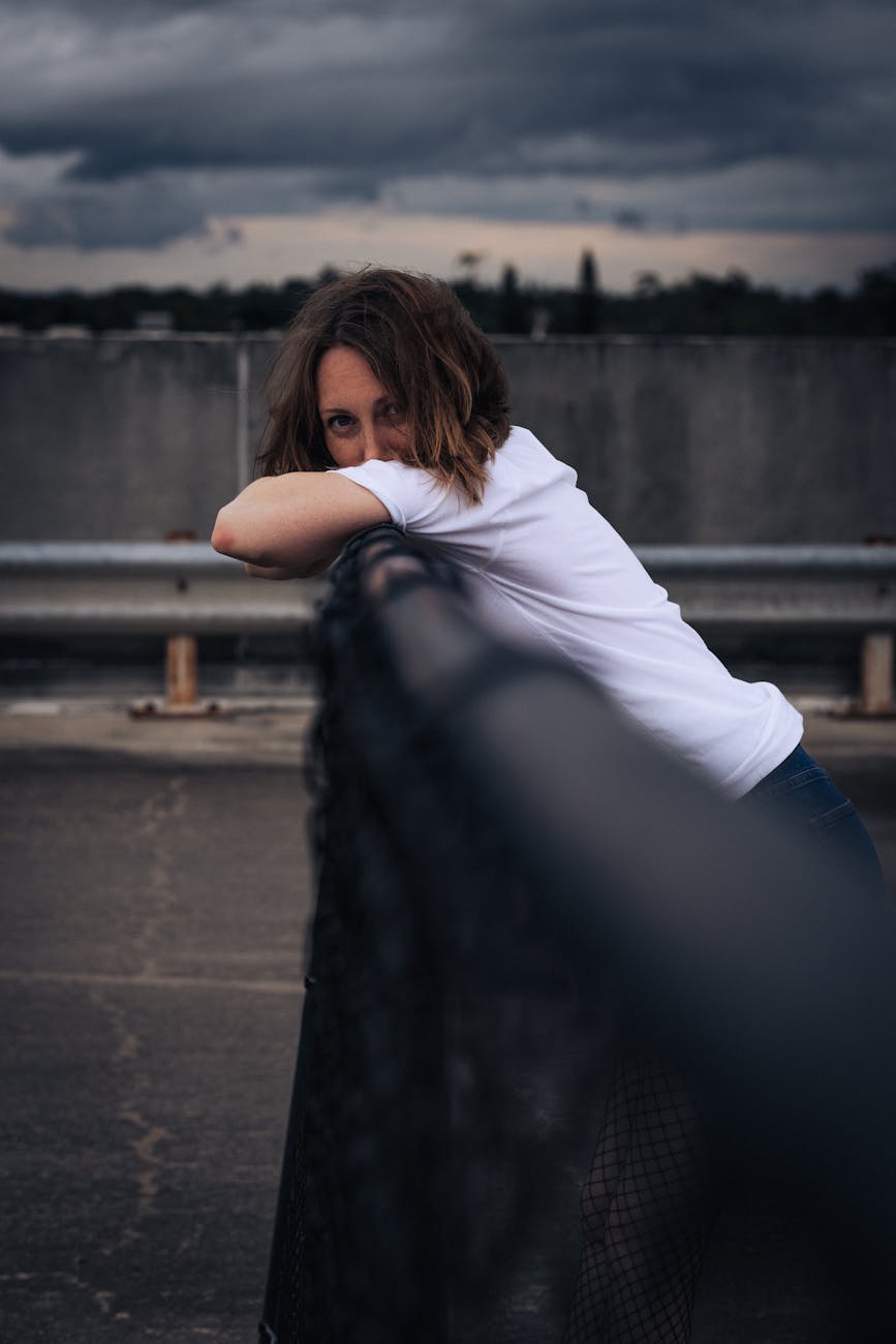 A woman leans on a railing under a cloudy sky in an urban setting, exuding a contemplative mood.