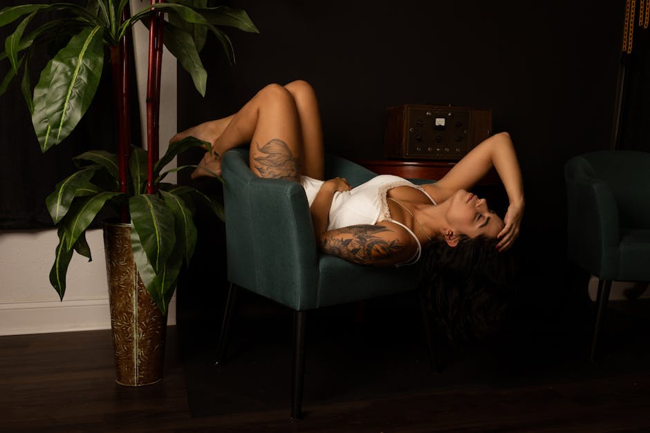 Woman with tattoos posing relaxed on a chair indoors beside a plant.