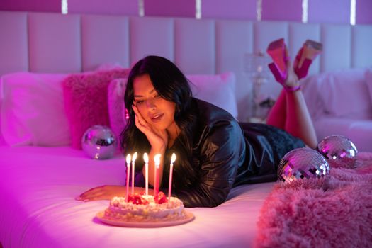 A woman lies on bed, admiring a birthday cake with lit candles, surrounded by disco balls.