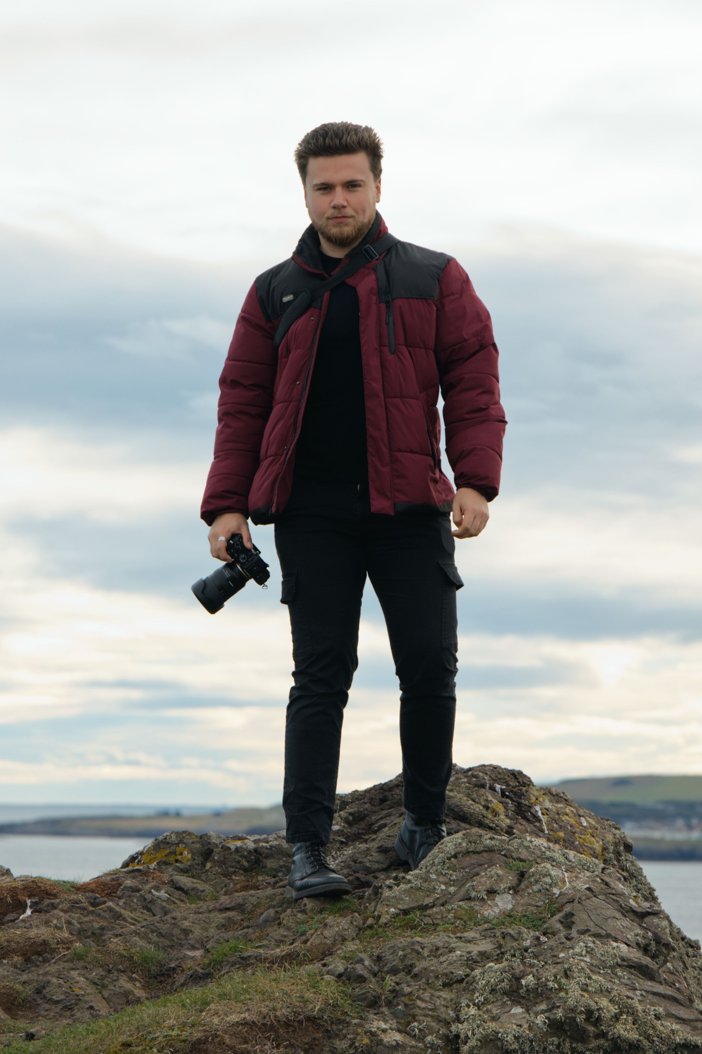 Male photographer holding a camera while standing on a rugged cliff edge against a cloudy sky.