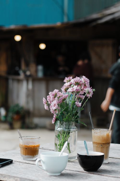 Free A serene cafe scene outdoors with coffee cups and a flower arrangement on a rustic table. Stock Photo