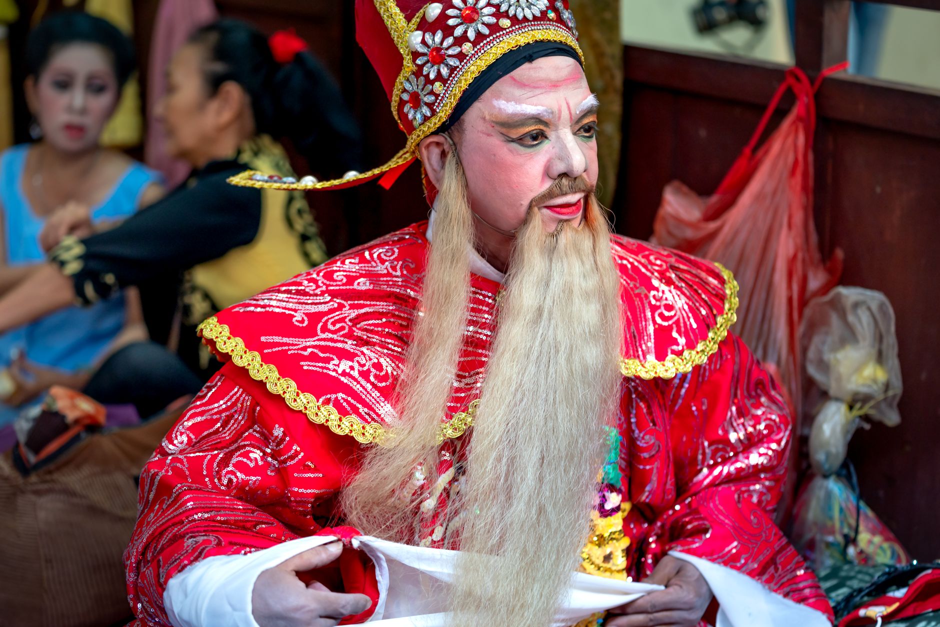 A Chinese opera performer in a red costume prepares backstage with detailed makeup.