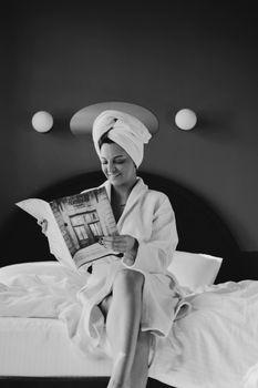 A woman in a bathrobe reads a magazine while sitting on a bed, exuding relaxation and tranquility.