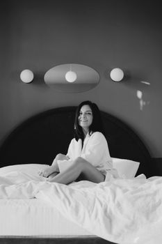 Monochrome portrait of a woman sitting on a bed, smiling confidently in a minimalist bedroom.