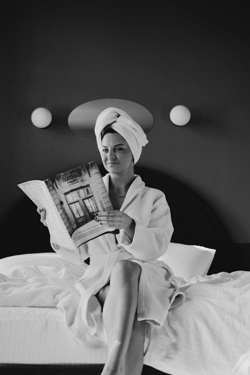 Grayscale Photo of a Woman Sitting in White Robe and Holding a Magazine