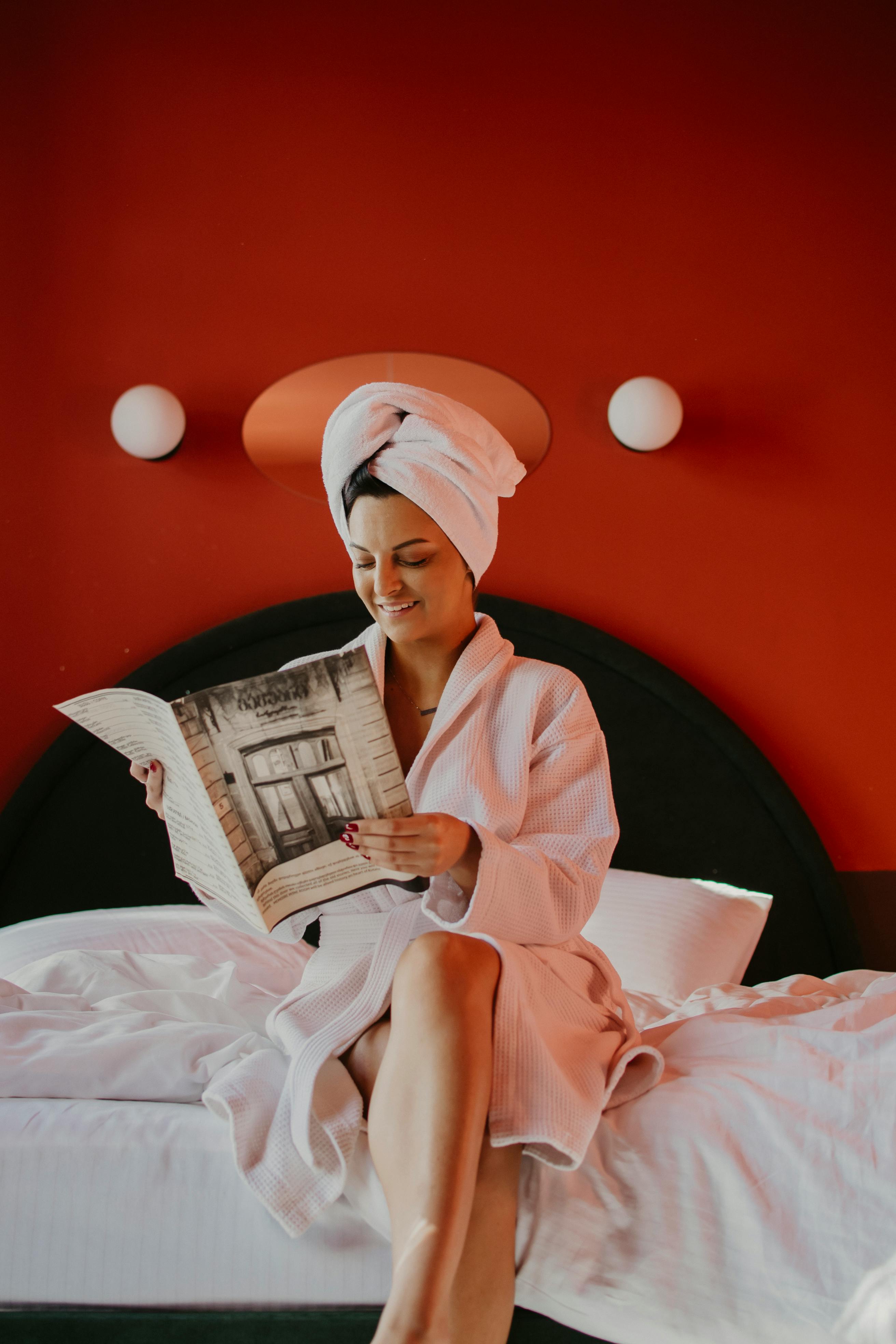 Woman in bathrobe reading on bed against red wall, exuding relaxation and modern interior design.