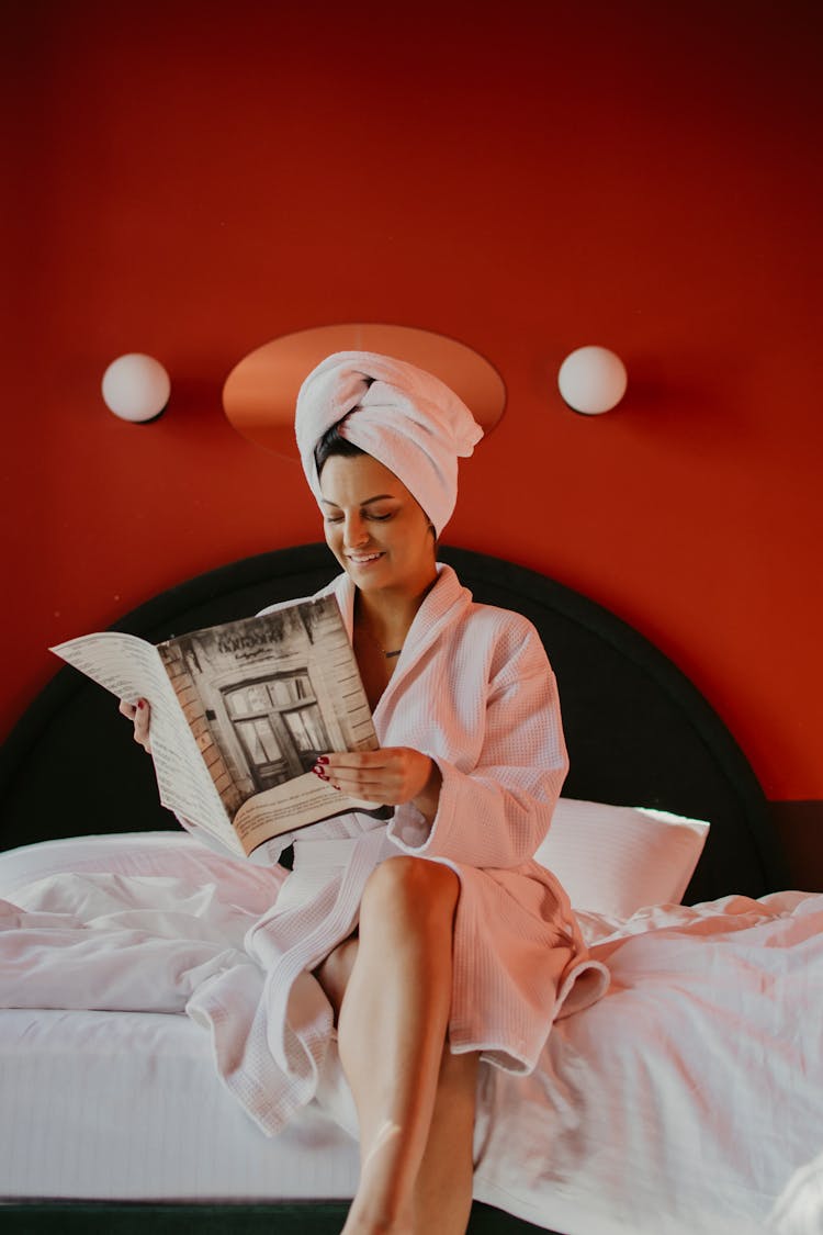 Smiling Woman In Bathrobes Sitting On Bed And Reading