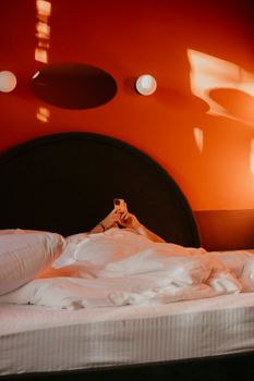 Individual lying in a hotel bed using a smartphone, set against distinctive orange wall decor.
