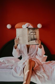 A woman relaxing in a hotel room, wrapped in a bathrobe, reading a magazine with a red wall backdrop.