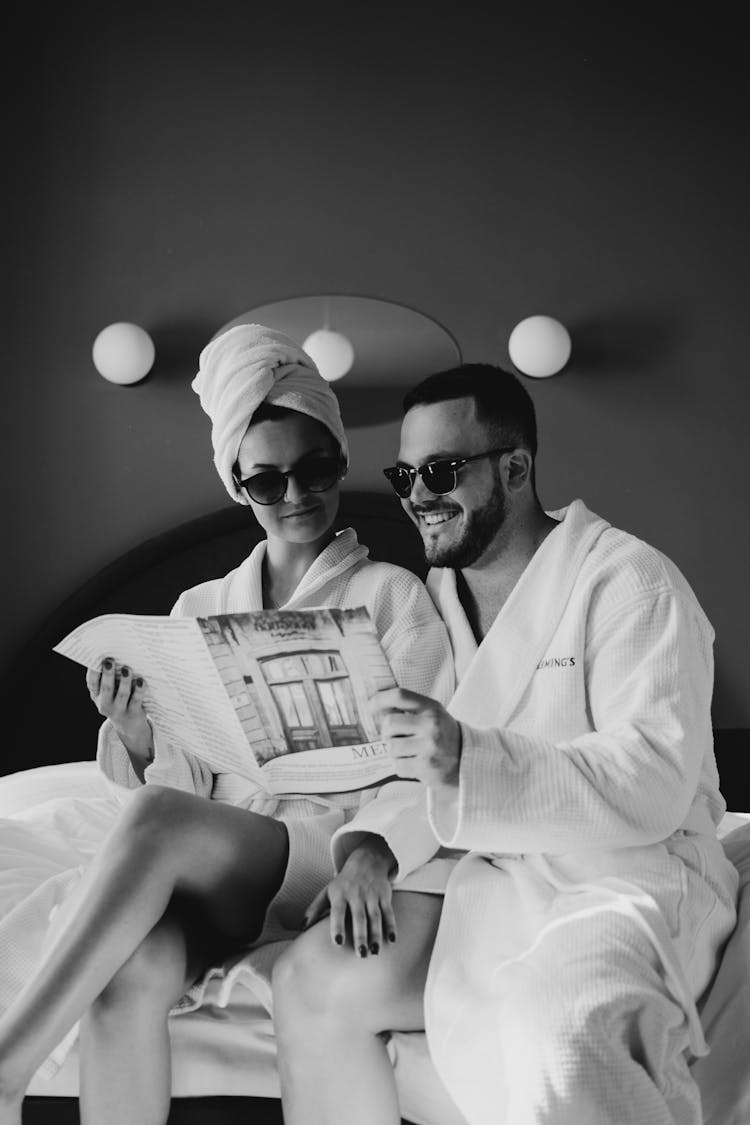 A Grayscale Of A Couple In Robes Reading A Magazine