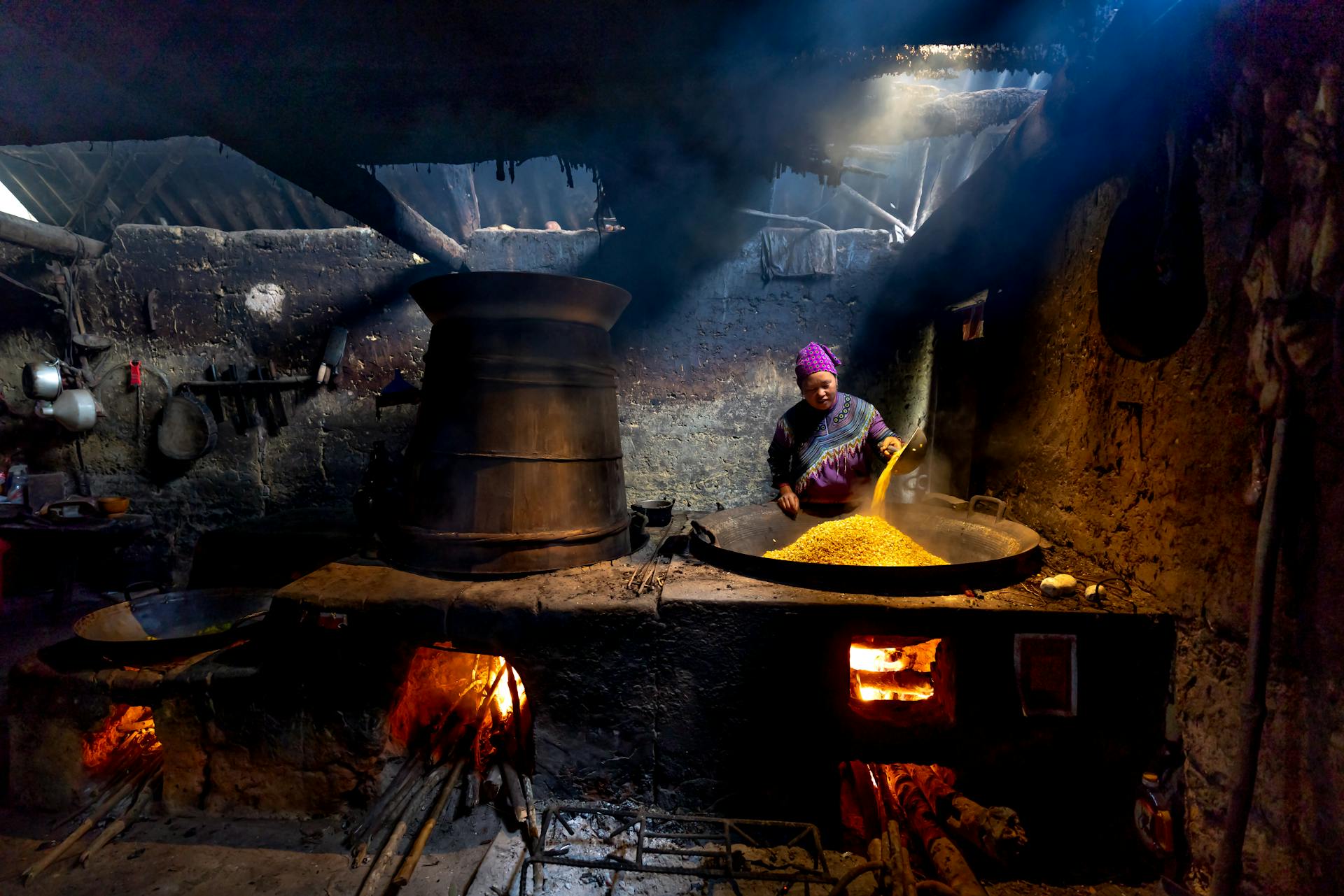 A woman cooking maize in a traditional rustic kitchen, with a furnace and fire.