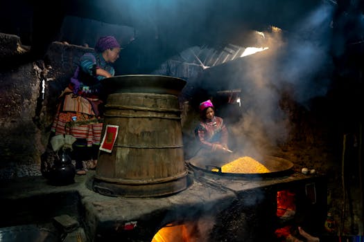 Two women prepare food in a rustic kitchen with traditional attire and large cooking pots.