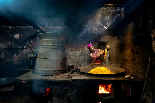 Asian woman cooking corn kernels over a smoky stove in a traditional kitchen.
