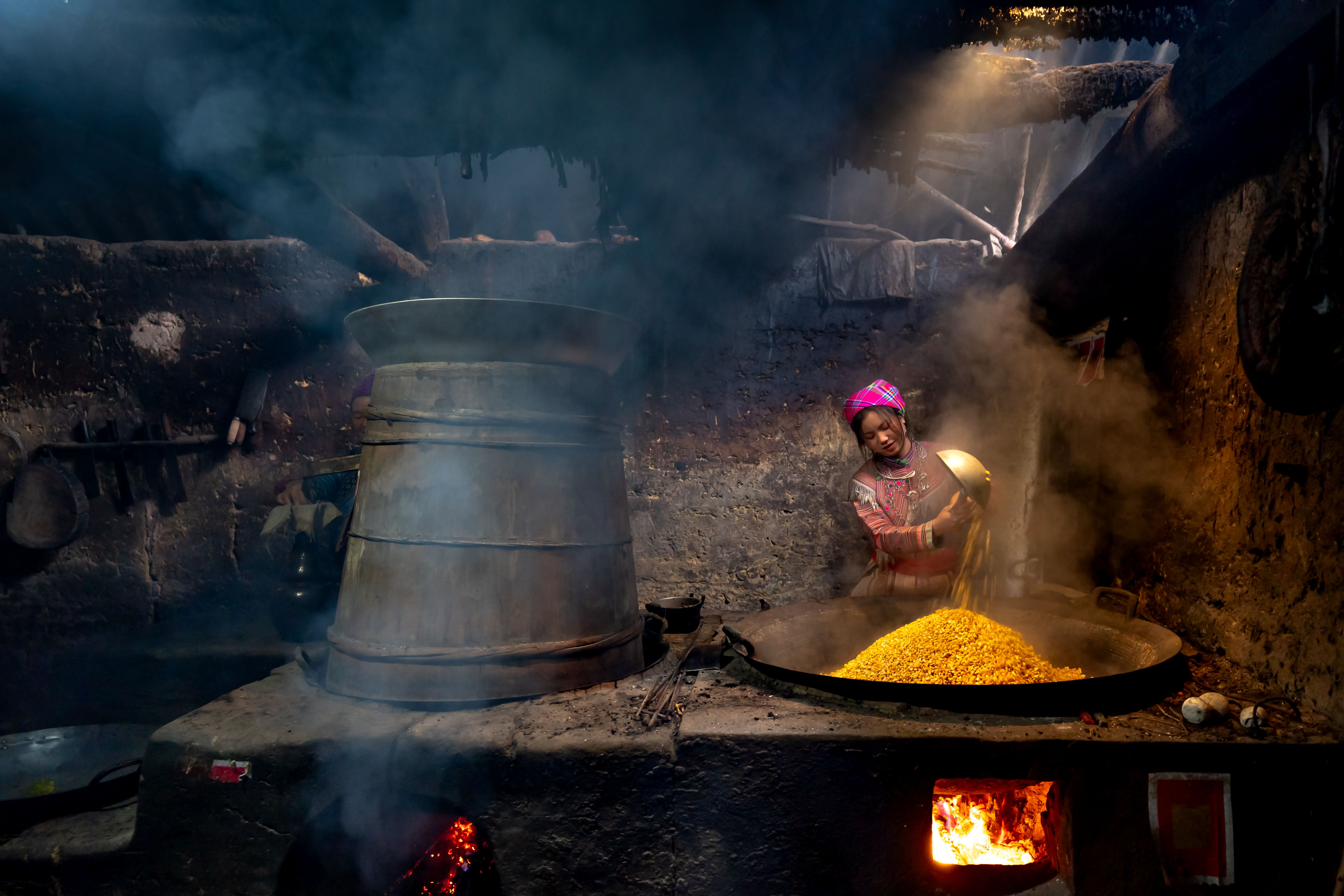 A woman stirring a large clay pot over an open fire at dawn, with steam rising and Mount Adaklu silhouetted in the background