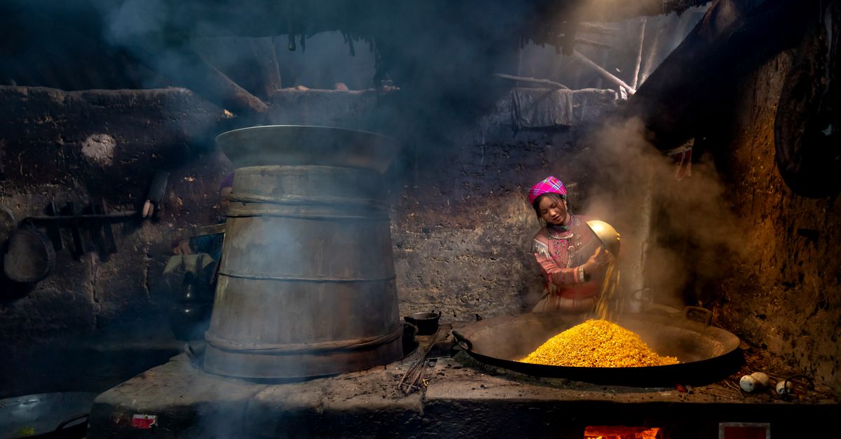 A woman stirring a large clay pot over an open fire at dawn, with steam rising and Mount Adaklu silhouetted in the background