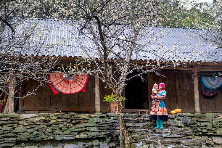 Woman In Traditional Costume With Child Near Wooden House