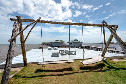 Free Rustic wooden swing overlooking seaside cabins on a sunny day. Perfect for a serene escape. Stock Photo