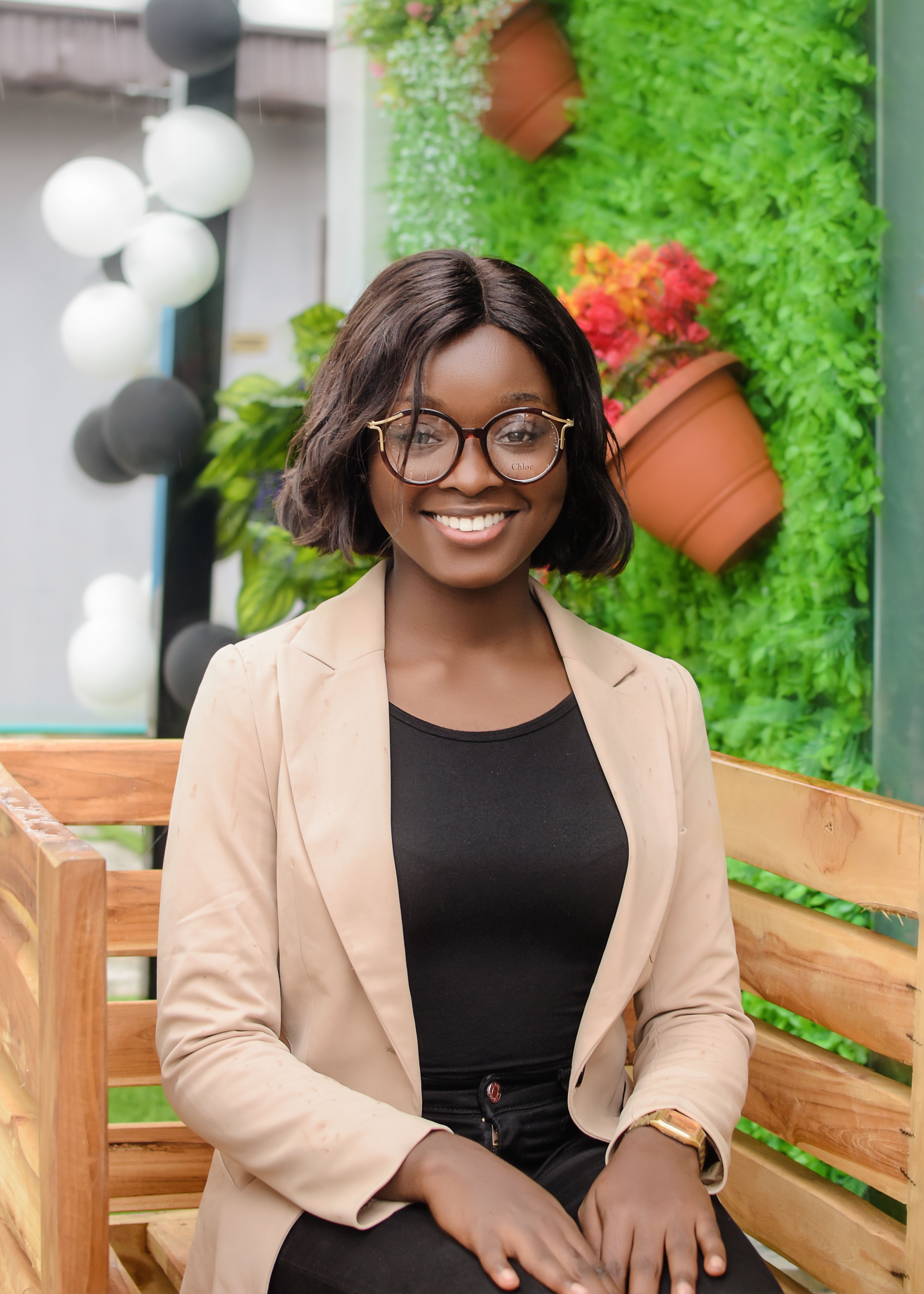 Smiling young woman wearing glasses and a blazer, sitting outdoors on a wooden bench.