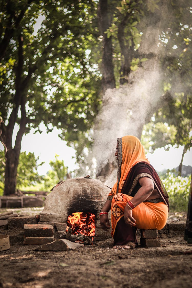 Old Woman In Traditional Clothes Sitting Near Fire In Forest