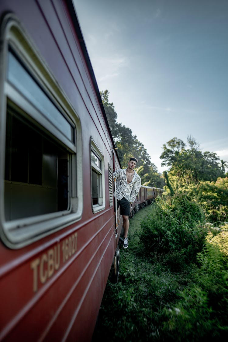 Man In Printed Shirt And Black Shorts Riding And Standing On The Door Of A Train