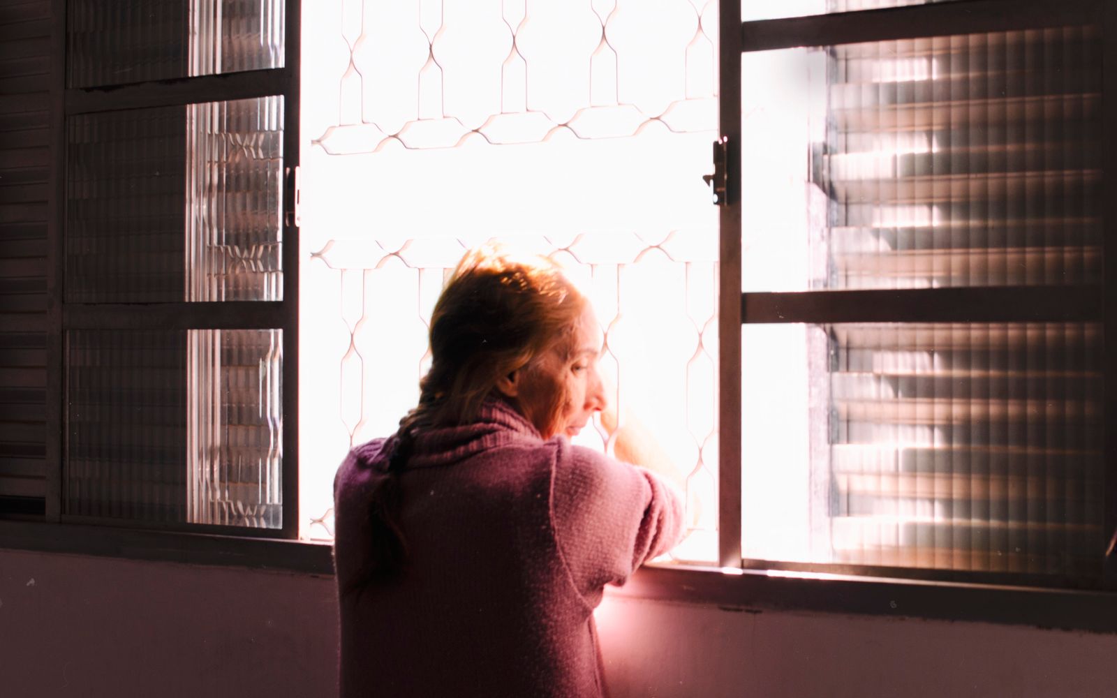 Woman in a soft pink sweater gazing out a sunlit window in contemplation