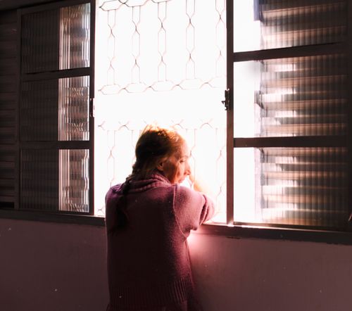 Free A senior woman looks through a window, bathed in soft sunlight, evoking contemplation. Stock Photo
